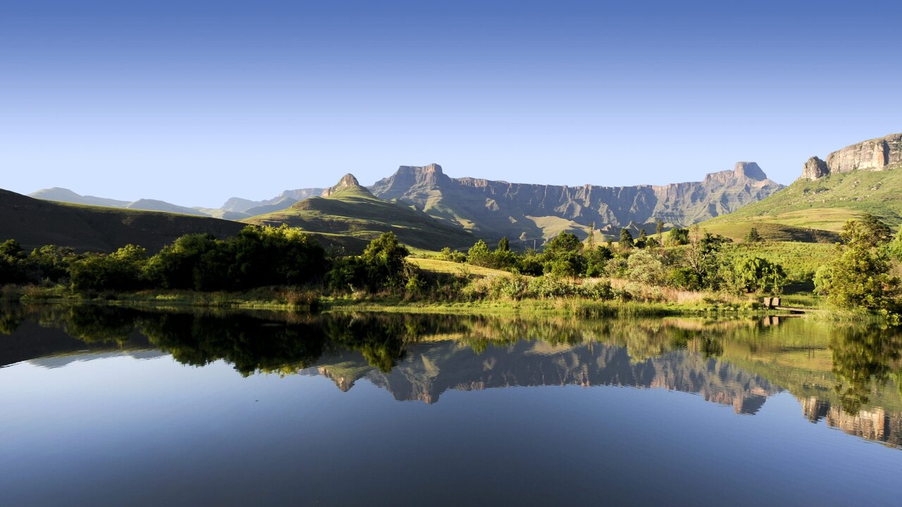 Um lago reflete colinas verdes e uma escarpa montanhosa recortada, com um pequeno cais de madeira na margem