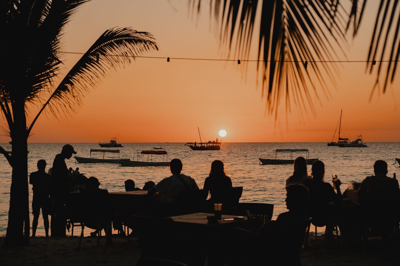 Pessoas num bar de praia sob palmeiras observam um dhow navegar ao entardecer
