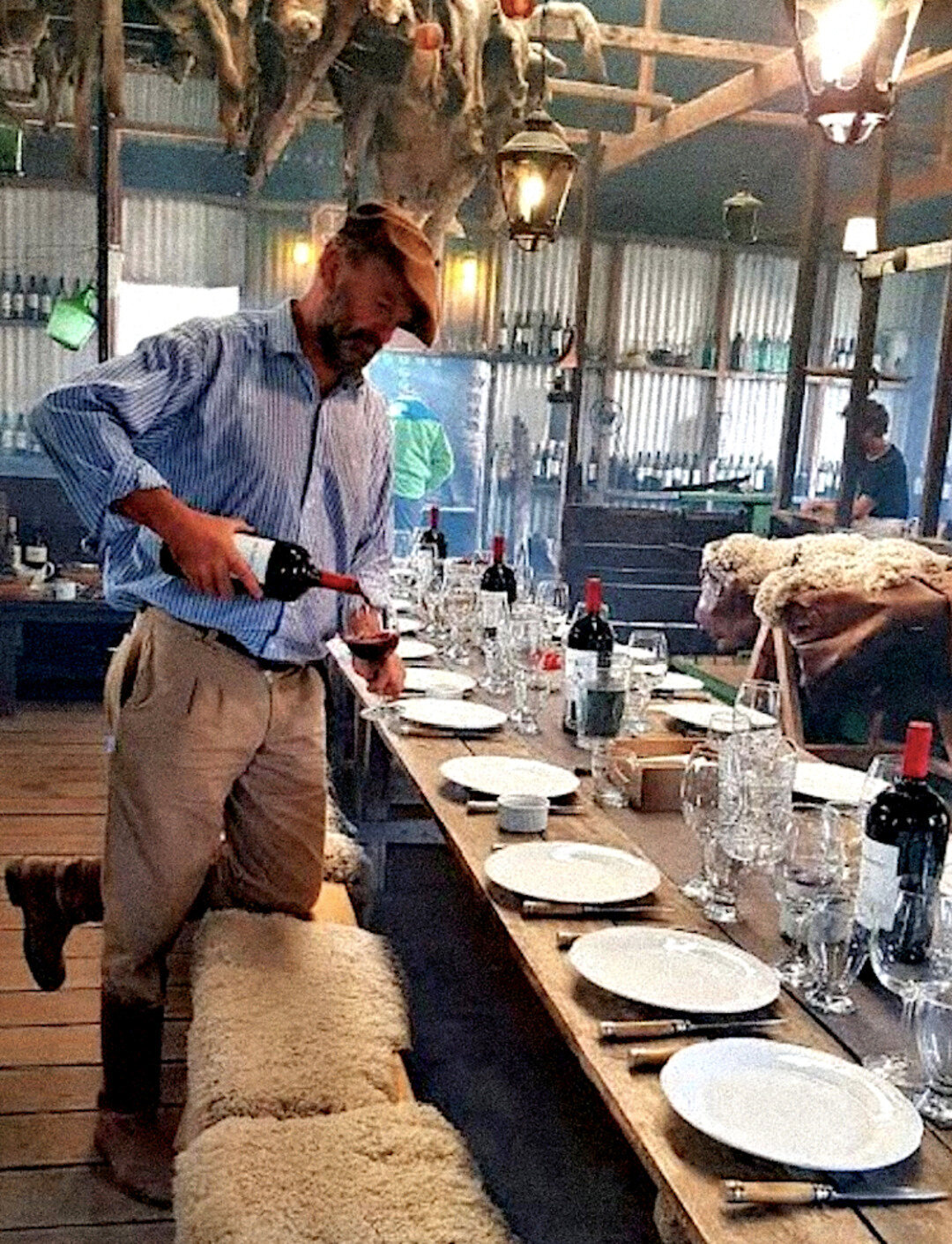 A man in a wide brimmed hat pouring red wine at a long table set for a group dinner in a rustic barn