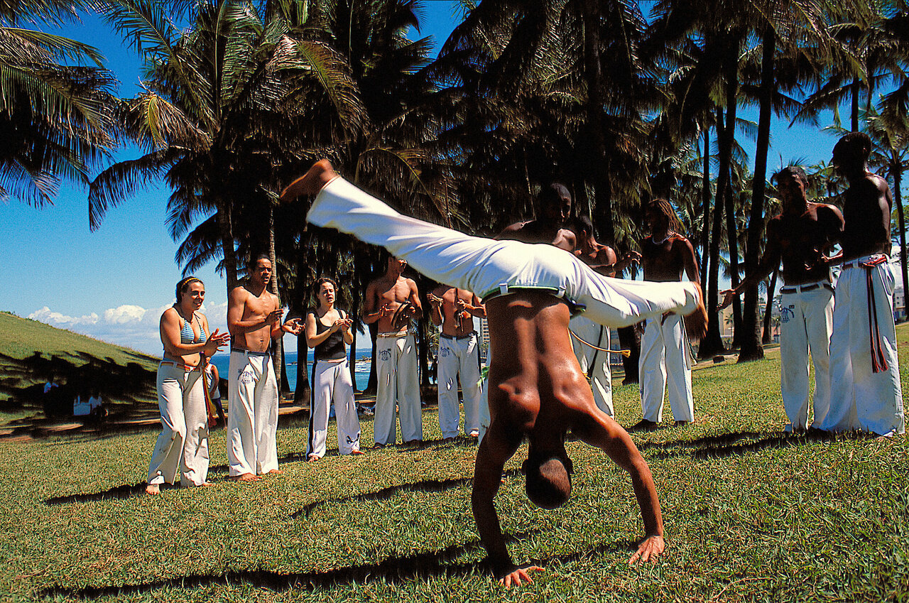Una exhibición de capoeira bajo las palmeras con espectadores aplaudiendo y la costa del océano al fondo