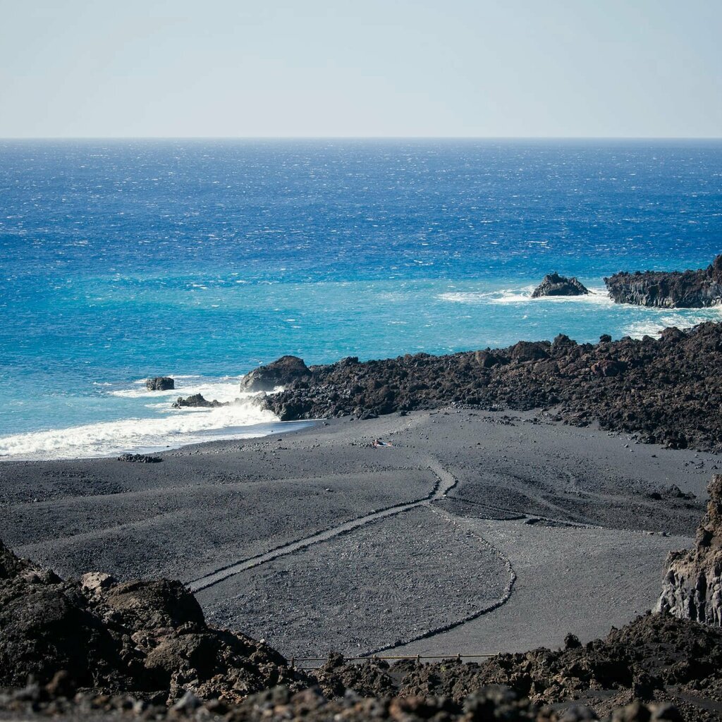 Costa rocosa volcánica con agua turquesa y un sendero de arena negra que lleva al mar